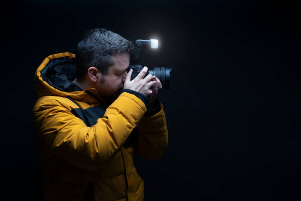 istock Photographer in a yellow coat taking a photo with flash with a black background 1553598267