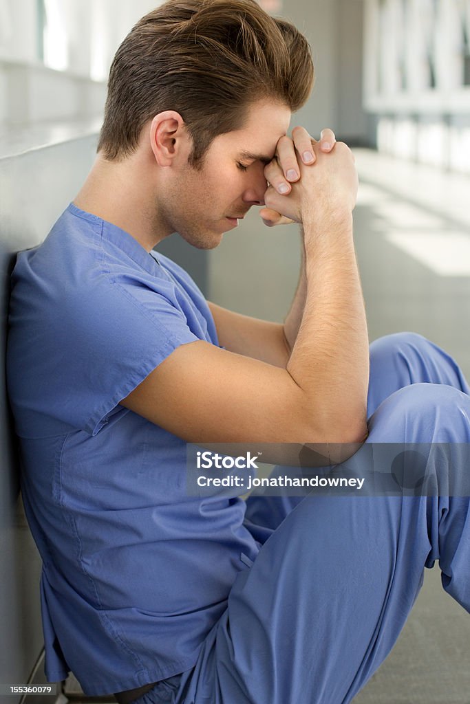 Young Man in Blue Scrubs A young man wearing blue scrubs sits with his head on his folded hands Male Nurse Stock Photo