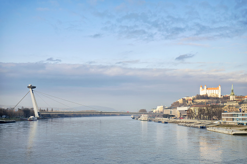 A view up the River Danube of the Slovakian Capital City, Bratislava, with the Novy Most Bridge ahead, and Bratislava Castle in bright sunlight to the right.