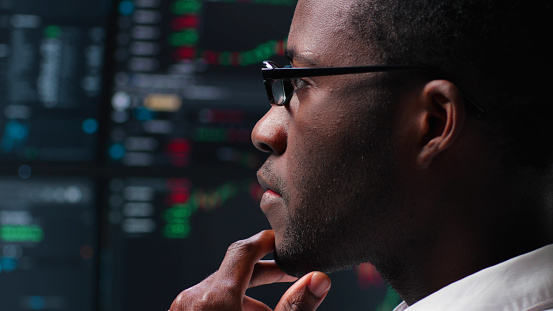 Close up of pensive African-American programmer looking at computer monitors with graphs. Trader analyze stock market data