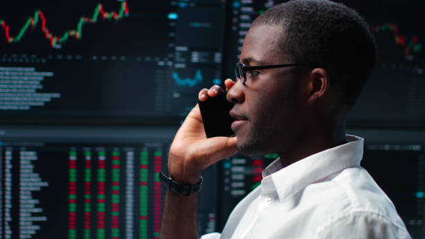 istock African-American trader talk on smartphone standing over multiple monitors with charts and data 1553630471