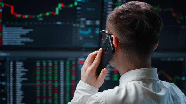 istock Back view of trader working with multiple computer screens full of charts and data talking on phone 1553634890