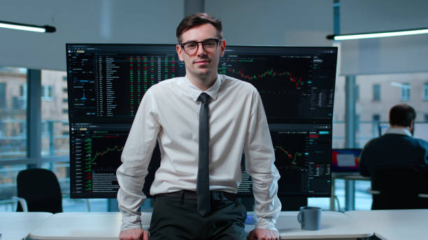 istock Portrait of happy trader looking at camera leaning on desk with multiple monitors 1553640521
