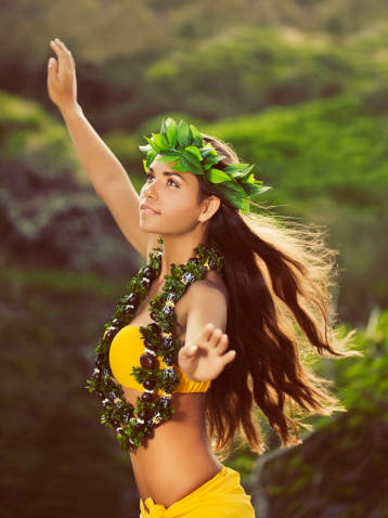 Young Hula Dancer posing for the camera with her hands in the air, wearing a lei and haku headpiece, shot from the waist up.