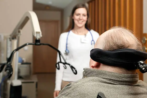 A home-care nurse approaches a patient seated in a power chair with headreast. The patient has advanced Multiple Sclerosis (MS) and has chosen to have his care done at home rather than a medical facility. The patient lift to the left will be used to transfer the man from his chair to bed. Today various aspects of bringing medical care to the home are gaining in popularity and this image depicts an advanced level of care.