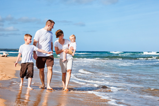 Photo of a young family of four enjoying a sunny day at  a Hawaiian beach, walking in the surf as they talk  to one another.