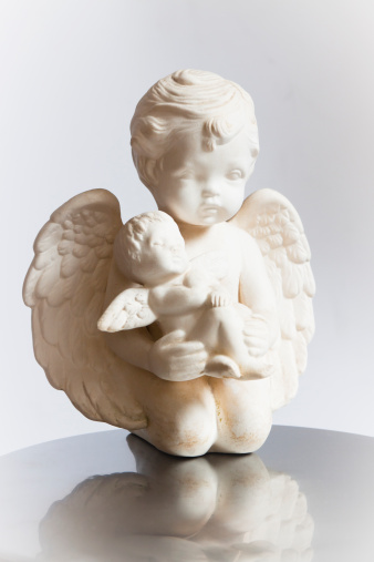 Small white statue of kneeling angel holding baby angel with reflection, full frame vertical composition with copy space