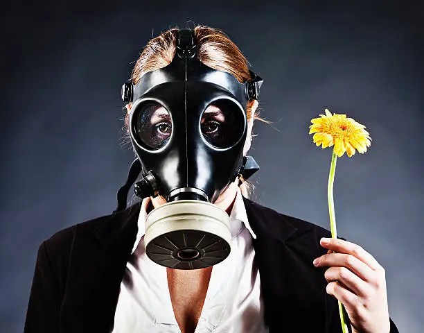 Sad-eyed woman in a gas mask holds a flower.She can't smell it but the need to wear the gasmask could be a pollution situation, war zone contamination or simply allergies or asthma! 