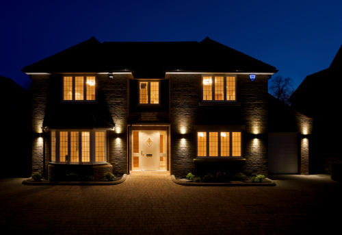 a view of the front and driveway of a lovely double fronted luxury new home, taken at dusk with all of the interior and exterior lights switched on. The branding on the alarm box has been changed to a fictional text.