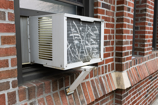 A closeup of a basic window air conditioner in the window of an old brick apartment building in Queens of New York City