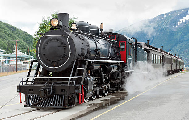 Steam train driving down tracks in Skagway, Alaska  The White Pass steam train waiting in the train station sidings at the port of Skagway with a cruise ship in the background road going steam engine stock pictures, royalty-free photos & images