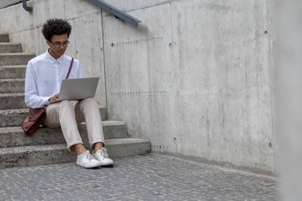 istock Curly-haired young man in white shirt sitting on the steps with laptop 1553726854