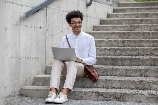 istock Curly-haired young man in white shirt sitting on the steps with laptop 1553726870
