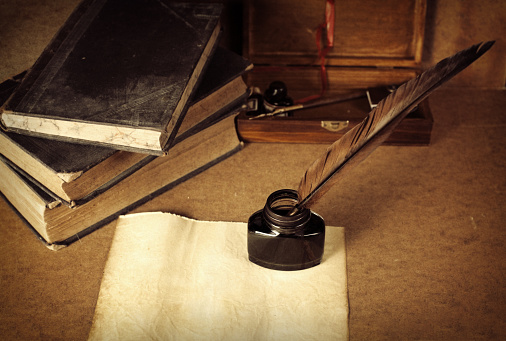 Antique desktop surface with old books, pen kit and blank parchment with quill pen in inkwell. Same series: