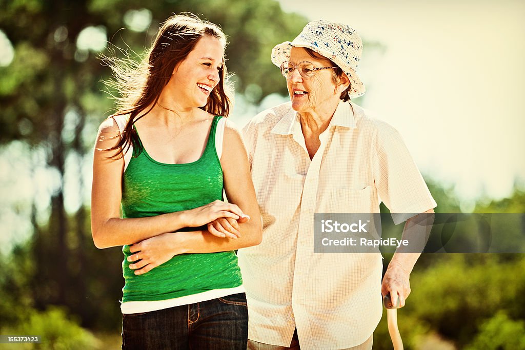 Granddaughter and grandmother smile as they go walking together A young woman and an old woman, bent over and using a stick, link arms and chat happily to each other laughing as they walk through a sunlit forest.  20-29 Years Stock Photo