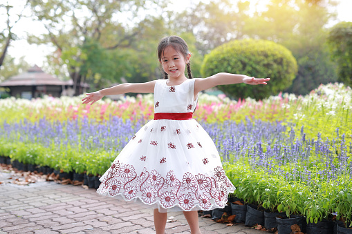 Happy Asian little child girl open arms wide and smiling in the fresh flower garden. Kid play in the park outdoor.