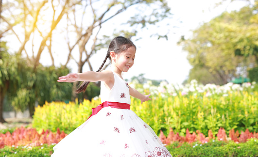 Asian little girl playful in flower garden with smiling in the sunny day. Kid open arms wide and play in nature park outdoor.