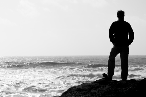 Black and white silhouette of a man standing on a rocky point looking at sea