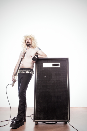A stereotypical 1980s style heavy metal rock and roll player leans on a large guitar amplifier cab.  The man is shirtless with gawdy boots and long curly blond hair, holding his white bass guitar.  Vertical with copy space.