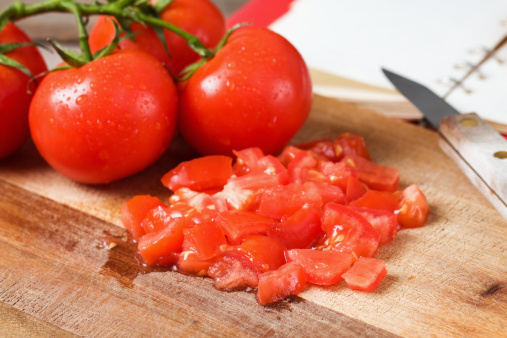 Chopped and whole vine ripened tomatoes on a wood cutting board with a kitchen knife and cook book.  Focus on the front chopped tomatoes.