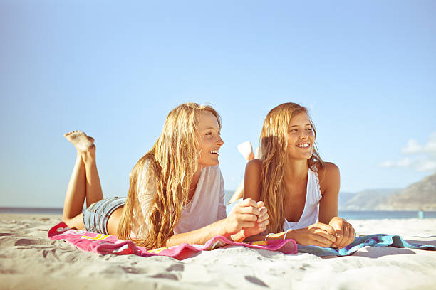 Two attractive teenage girls relaxing and gossiping on the beach Two teenage South African girls gossiping and laughing together while they sunbathe on Long Beach in Kommetjie (just outside of Cape Town), South Africa. girls laying on beach stock pictures, royalty-free photos & images