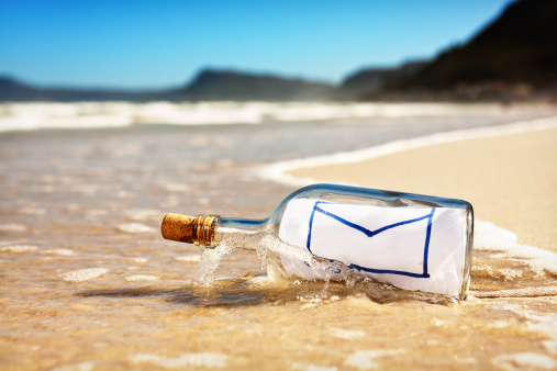 At water's edge on a beautiful but deserted beach lies a bottle containing a blank hand-drawn envelope, the universal icon for email. 