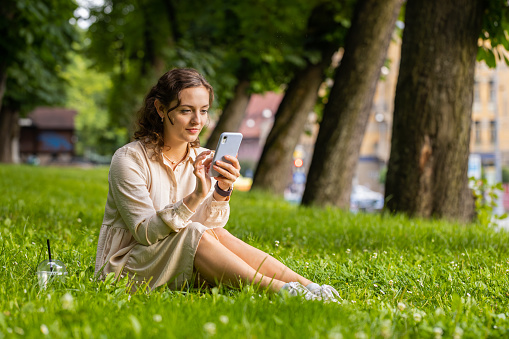 Smiling young woman using smartphone typing text answering messages chatting online looking mobile screen social media app. Girl sitting on grass in urban sunset city park holding phone in hands