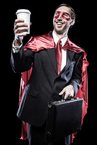 A business man in suit and tie wears a superhero cape and mask as well, smiling with his to go paper coffee cup and briefcase.  Isolated on black background.