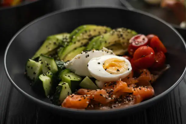 Salad of salmon, cucumber, tomato, egg, avocado and sesame seeds on a black plate, close-up. Vegan restaurant menu. Copy space