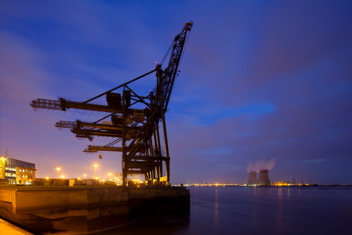 A container terminal with tall cranes with a nuclear power station in the background. Night shot taken in Antwerp, Belgium.
