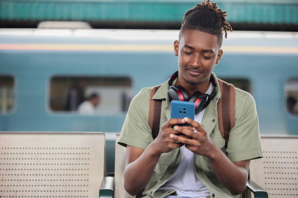 istock Young Man Commuting To Work On Train Sitting  On Platform Looking At Mobile Phone For Travel Information Or Social Media 1553801556