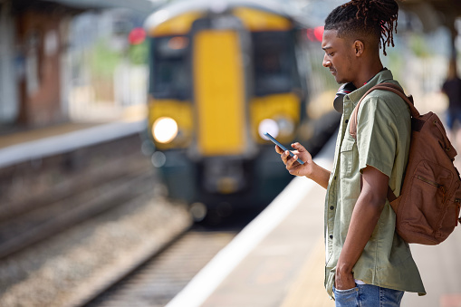 Young Man Commuting To Work On Train Standing  On Platform Looking At Mobile Phone As Train Arrives