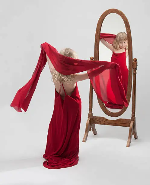 A little blond girl playing dress-up in an oversize gown and admiring her reflection in a full length dressing mirror. close pin holding gown together.