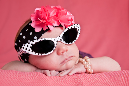 An adorable 1 month old baby girl dressed in sunglasses, headband and pearl bracelet.