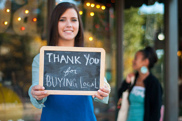Thank you for buying local Young business owner holding a "Thank You for Buying Local" sign in front of her store with shopper in the background. locol shopping stock pictures, royalty-free photos & images