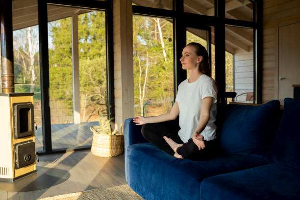istock Cheerful young lady is sitting at home on a comfortable blue sofa and meditating. A woman does yoga in the lotus position on the sofa, finds inner balance, copes with stress indoors 1553829443