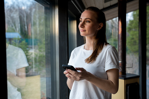 Smiling young woman using a smartphone, standing at the window of the house, looking at the phone screen, chatting or shopping online.