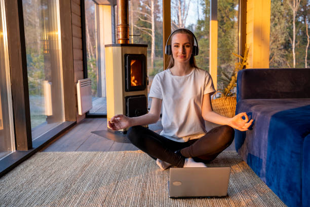 Young woman wearing headphones during an online yoga meditation practice. A woman sits in a lotus position in front of a laptop monitor and meditates with her eyes closed in the living room against the background of a fireplace. mujer joven con auriculares durante una práctica de meditación de yoga en línea. una mujer se sienta en posición de loto frente a un monitor de computadora portátil y medita con los ojos cerrados en la sala de estar contra el fondo de una chimenea.