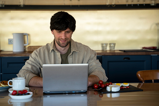 Smiling young man uses a laptop and has breakfast in the kitchen.