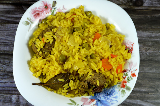 A plate of hot steamed long grain yellow Basmati rice with vegetables peas and carrots with sliced onion, bay laurel, cardamom and pieces of hot boiled beef meat isolated on wooden background, selective focus