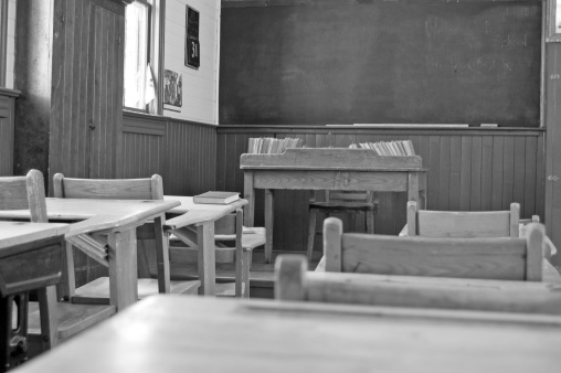 A black and white image of an early 1900's one room school with textbooks and the teacher's desk.