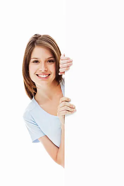 Beautiful young woman looks around a corner and smiles at the camera. Vertical shot. Isolated on white.