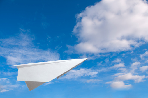 White paper plane against blue sky with fluffy clouds.