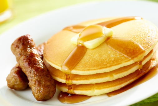 Tight shot of a stack of pancakes with pat of melted butter and syrup on top with sausage; OJ and Coffee cup in background. Professionally shot, color corrected, exported 16 bit and retouched for maximum image quality.