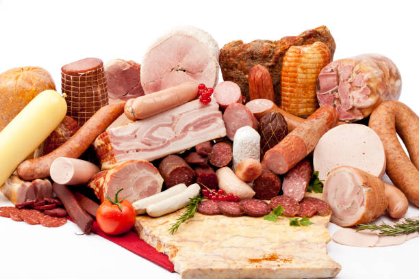 A Pile of meat featuring smoked meats, bacon, salami, baloney, sausages, kielbasa, pork roll and some herbs and a tomato.