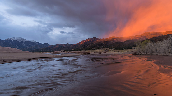 A ray of twilight illuminates misty clouds hovering over rolling hills and rushing Medano Creek on a stormy Spring evening. Great Sand Dunes National Park, Colorado, USA.