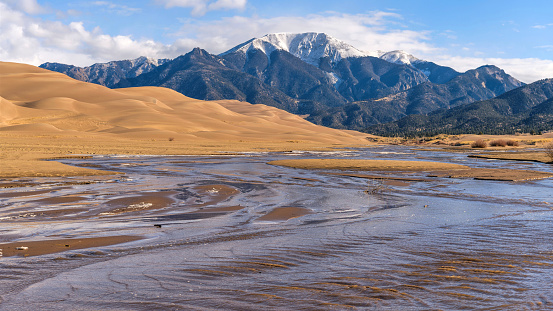 A bright sunny Spring morning view of Medano Creek rushing down a sandy valley at base of rolling Great Sand Dunes and snow-capped Mt. Herard. Great Sand Dunes National Park, Colorado, USA.