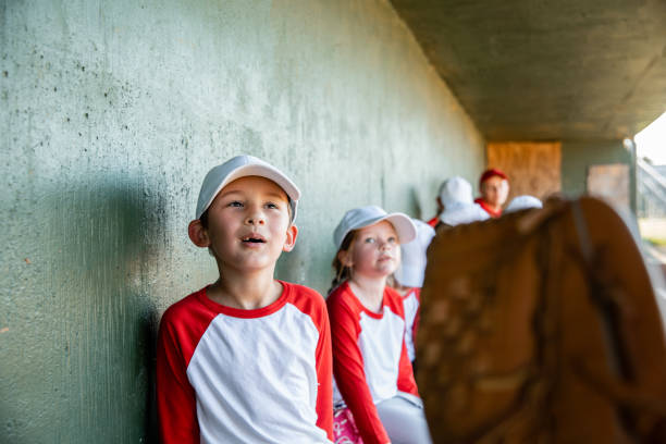 リトルリーグの野球の試合中にチームメイトとダッグアウトに座る小さな男の子 - dugout baseball bench bat ストックフォトと画像