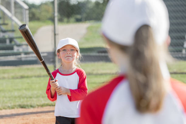 adorable jugador de ligas pequeñas sostiene un bate de béisbol mientras juega con sus compañeros de equipo - baseball bat baseball little league baseballs fotografías e imágenes de stock