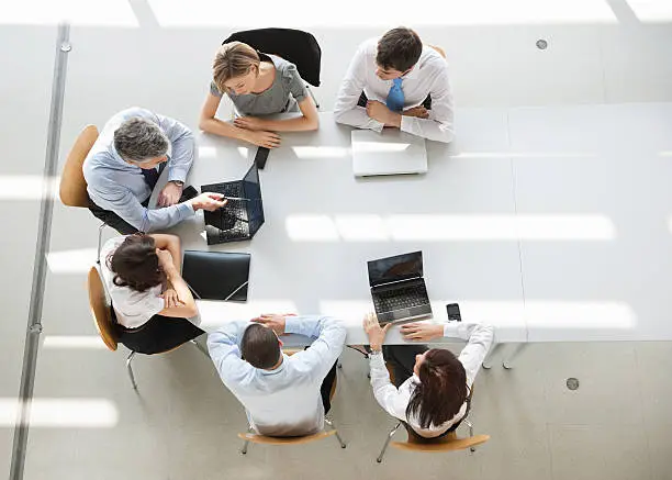 Business People In Meeting. A group of six men and women (three man and three woman) sitting around an oval white table having a business meeting. They all are dressed in business causal, and have their laptops out, only two are being used. They are sitting in simple wooden chairs. 